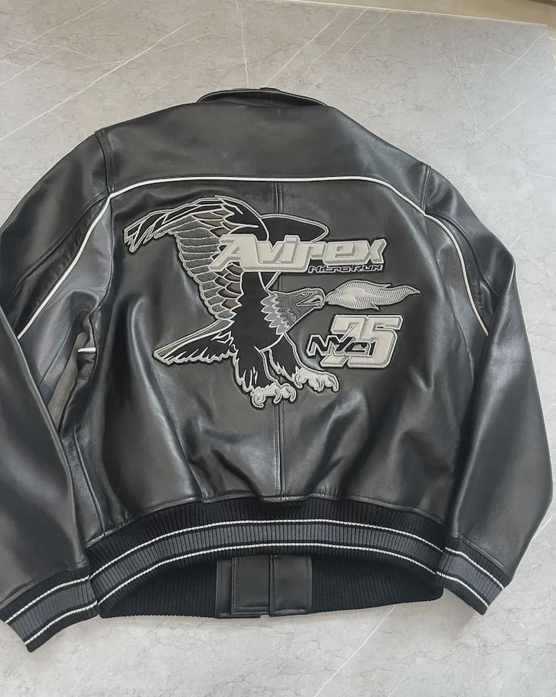 May include: Black leather jacket with an embroidered eagle design on the back. The jacket displays the text "Avirex History NYC 75" in silver and white. It has a ribbed hem and cuffs with white stripes.
