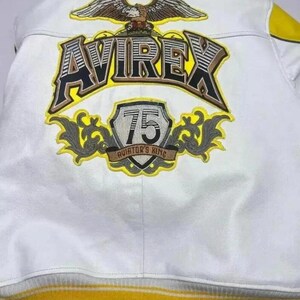May include: White leather jacket with yellow and grey accents. Features the brand name "AVIREX" in large, bold lettering, along with an eagle emblem and a "75 Aviator's King" patch. The jacket has a ribbed yellow hem.