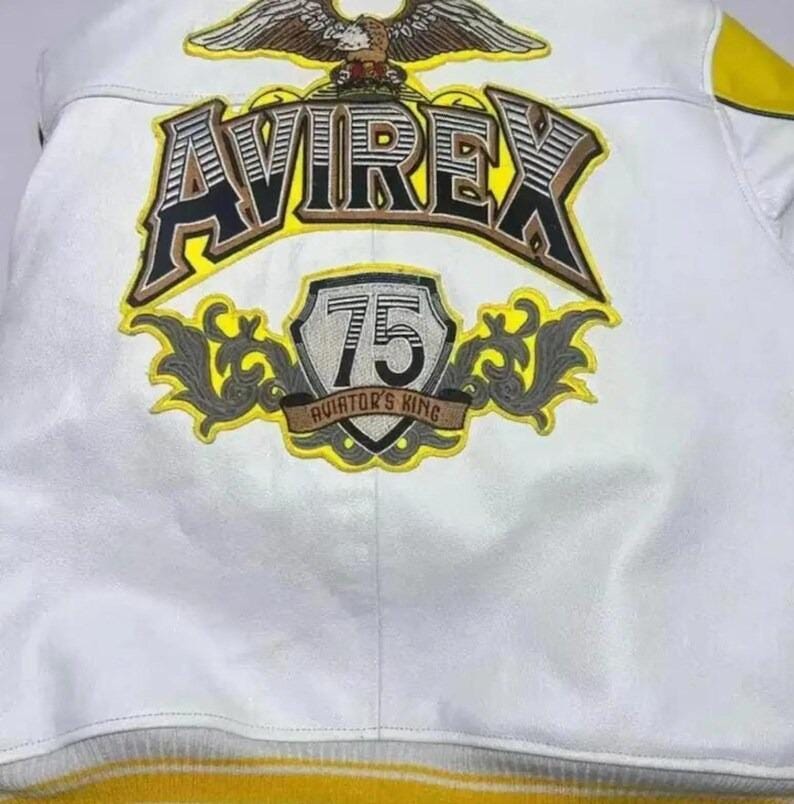 May include: White leather jacket with yellow and grey accents. Features the brand name "AVIREX" in large, bold lettering, along with an eagle emblem and a "75 Aviator's King" patch. The jacket has a ribbed yellow hem.