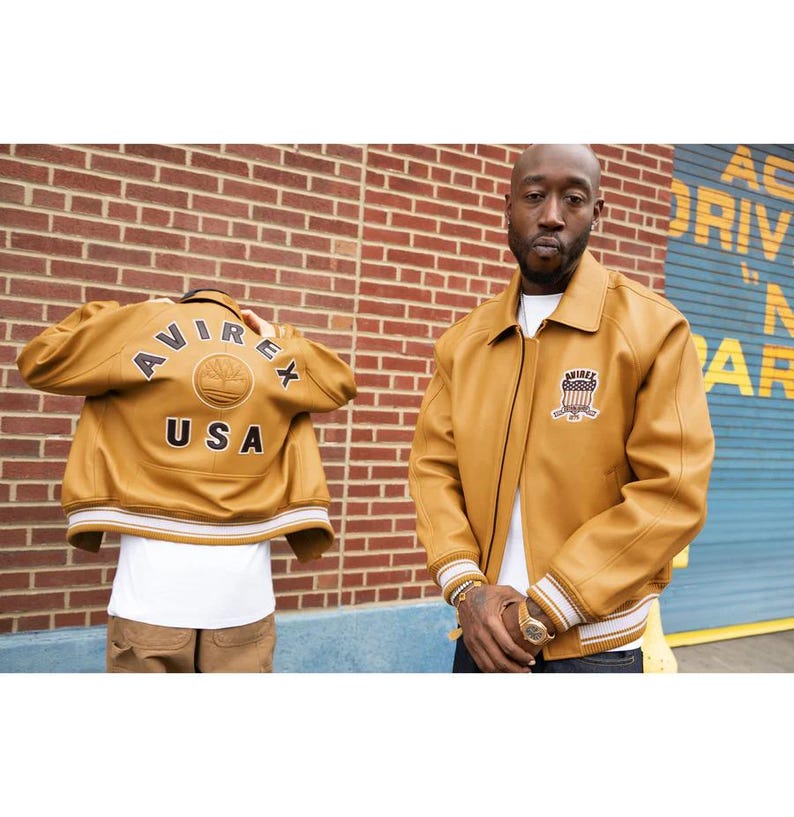 May include: Mustard-coloured leather jacket with a collared neckline and a patch on the chest. The back of the jacket features the text "AVIREX USA" in black lettering. The jacket has white and gold striped cuffs and hem.