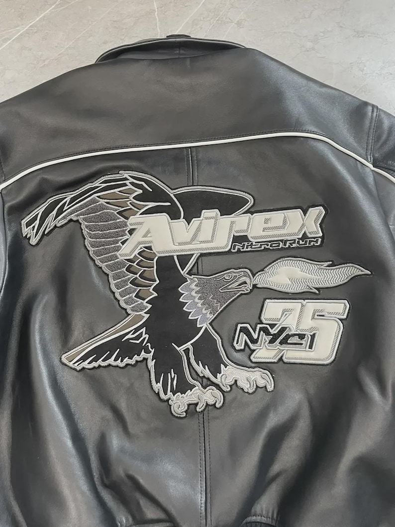May include: Black leather jacket featuring an embroidered eagle motif on the back. The design includes the text "Avirex Nitro Run" and "75 NYC". The jacket has white piping along the seams.