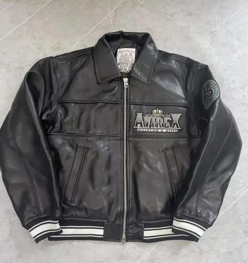 May include: Black leather jacket with a zip-up front, collar, and white striped cuffs. The jacket features the "AVIREX" logo in silver lettering on the chest and a patch on the sleeve. A classic bomber style.