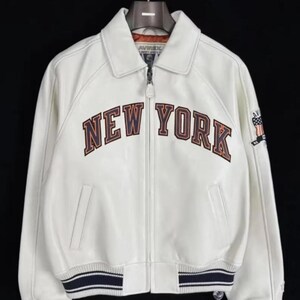 Pode incluir: Casaco de couro branco com o texto "NEW YORK" em letras marrons e laranja no peito. O casaco tem fecho de correr, gola e punhos e bainha listrados em azul marinho e branco. Um pequeno patch est&aacute; na manga direita.