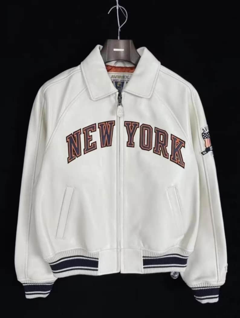 Pode incluir: Casaco de couro branco com o texto "NEW YORK" em letras marrons e laranja no peito. O casaco tem fecho de correr, gola e punhos e bainha listrados em azul marinho e branco. Um pequeno patch est&aacute; na manga direita.