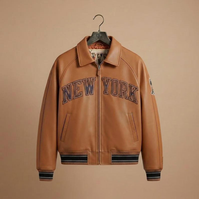 Puede incluir: Una chaqueta de cuero color canela con cremallera frontal completa, cuello cl&aacute;sico y pu&ntilde;os y dobladillo acanalados con rayas blancas y negras. El texto NEW YORK est&aacute; bordado en azul oscuro en el pecho y tiene un peque&ntilde;o parche en la manga.