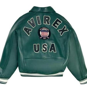 Może przedstawiać: Ciemnozielona sk&oacute;rzana kurtka z białymi wykończeniami. Z tyłu napis "AVIREX" w łuku, poniżej "USA" oraz okrągła naszywka z amerykańską flagą. Klasyczny styl bomberki.