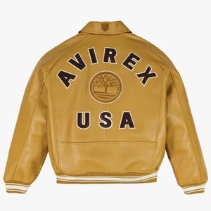May include: Mustard-coloured leather jacket with the text "AVIREX USA" in brown lettering. The jacket has a collared neckline, a ribbed hem with white stripes, and a logo patch on the upper back. The jacket is a classic bomber style.