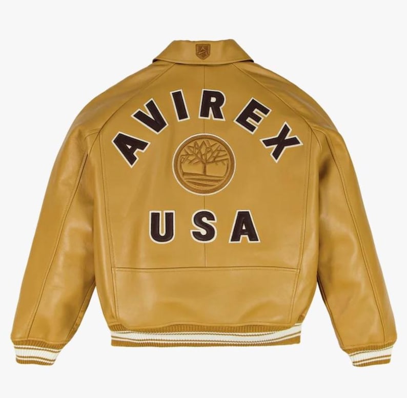 May include: Mustard-coloured leather jacket with the text "AVIREX USA" in brown lettering. The jacket has a collared neckline, a ribbed hem with white stripes, and a logo patch on the upper back. The jacket is a classic bomber style.