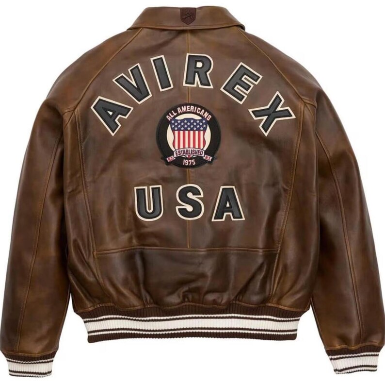 May include: Brown leather jacket with the word "AVIREX" across the shoulders and "USA" at the bottom. A circular patch with the American flag and the words "ALL AMERICANS ESTABLISHED 1975" is centered. The jacket has a ribbed white and brown striped hem.