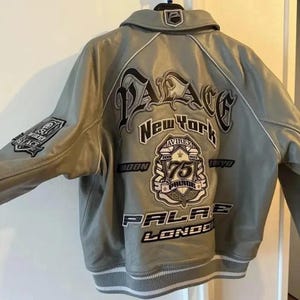 May include: A gray leather jacket with white trim and multiple patches. The back features the word "PALACE" above "NEW YORK" and a patch with "AVIREX 75". Additional patches include "PALACE LONDON" and "MOON".