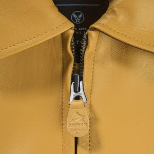 May include: Close-up of a mustard-coloured leather jacket with a silver zip. The zip pull features the brand name "AVIREX" and a logo. The jacket's collar is visible, and the lining has text that reads "CIVILIAN & MILITARY TAILORS".