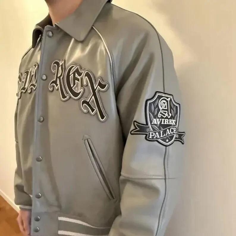 May include: A gray leather jacket with the text "REX" in a black and white font on the front. The jacket has a patch on the sleeve that says "AVIREX PALACE". The jacket has a button-up front and white trim.
