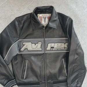 May include: Black leather jacket featuring the text "Avi Rex" in silver and grey across the chest. The jacket has a zip-up front, two front pockets, and a ribbed hem and cuffs. The collar has a small tag with text.