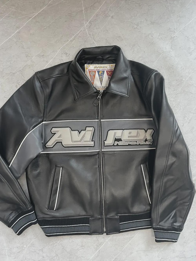 May include: Black leather jacket featuring the text "Avi Rex" in silver and grey across the chest. The jacket has a zip-up front, two front pockets, and a ribbed hem and cuffs. The collar has a small tag with text.