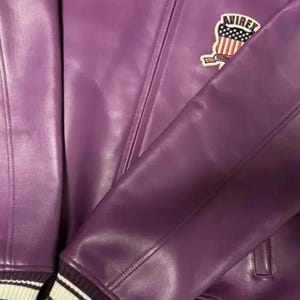 Puede incluir: Primer plano de una chaqueta de cuero morada con un pu&ntilde;o a rayas blancas y negras. La chaqueta presenta un parche con el texto "AVIREX" y un dise&ntilde;o de estrellas y rayas.