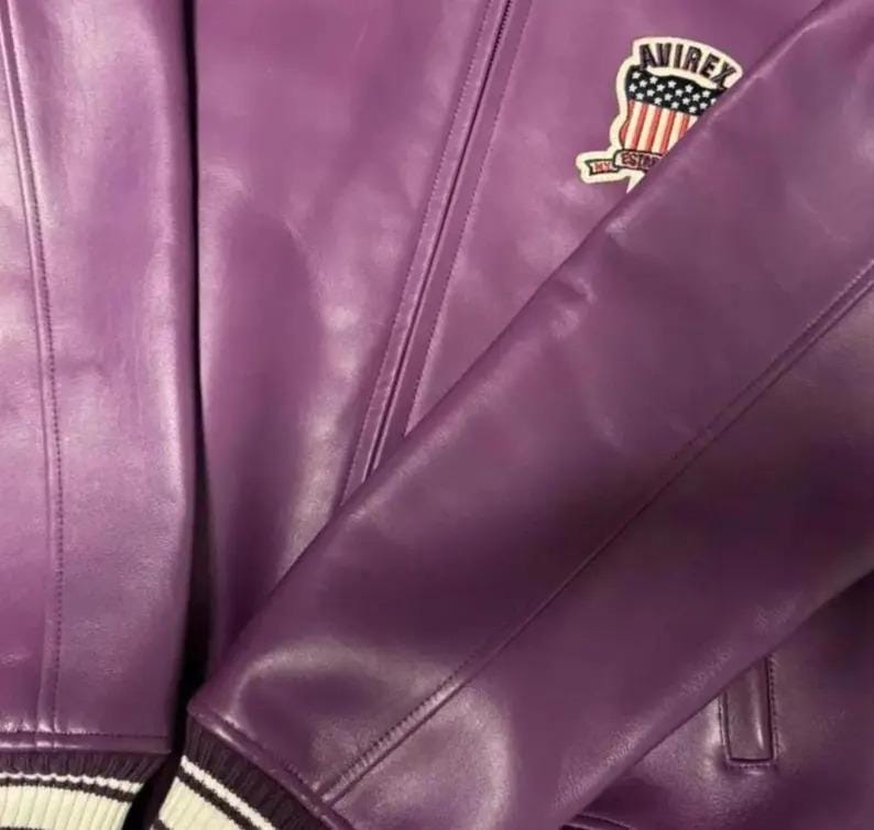 Puede incluir: Primer plano de una chaqueta de cuero morada con un pu&ntilde;o a rayas blancas y negras. La chaqueta presenta un parche con el texto "AVIREX" y un dise&ntilde;o de estrellas y rayas.