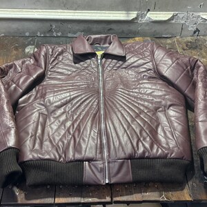 May include: A burgundy leather jacket with a quilted design and a zipper closure. The jacket features a ribbed collar and cuffs, and decorative stitching on the shoulders and chest. The jacket is a medium size.
