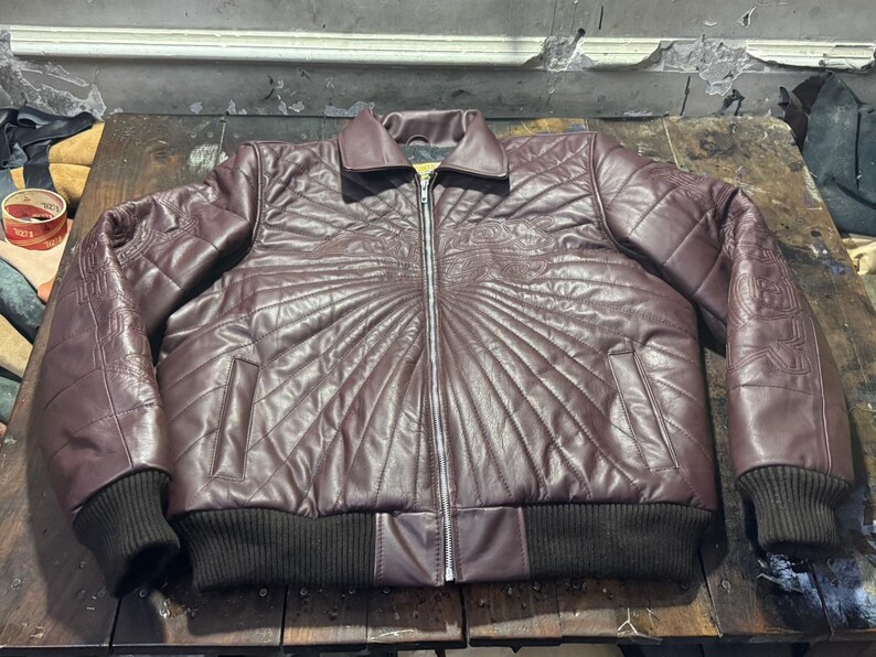 May include: A burgundy leather jacket with a quilted design and a zipper closure. The jacket features a ribbed collar and cuffs, and decorative stitching on the shoulders and chest. The jacket is a medium size.