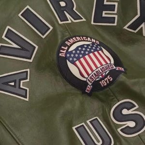 May include: Close-up of a green leather jacket with the word "AVIREX" in large, black, block letters outlined in white. A patch featuring the American flag and the words "ALL AMERICANS ESTABLISHED 1975" is also visible.