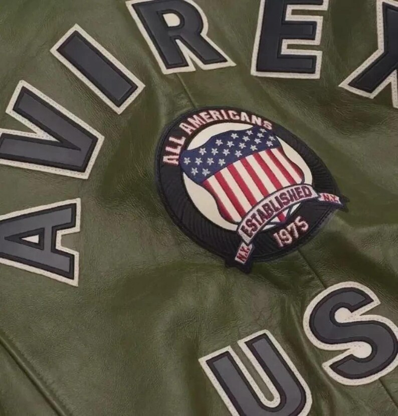 May include: Close-up of a green leather jacket with the word "AVIREX" in large, black, block letters outlined in white. A patch featuring the American flag and the words "ALL AMERICANS ESTABLISHED 1975" is also visible.