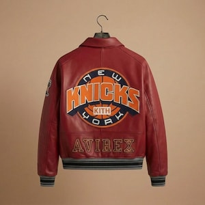 Puede incluir: Una chaqueta universitaria de cuero rojo con un gran parche circular en la espalda con el texto NEW YORK KNICKS y KITH en naranja y azul marino. La palabra AVIREX est&aacute; bordada en dorado debajo del parche. La chaqueta tiene pu&ntilde;os acanalados.