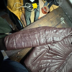 May include: A burgundy leather jacket with quilted stitching and a ribbed cuff. The jacket features the word "AMOR" stitched on the sleeve. The jacket has a front pocket and a zipper closure.