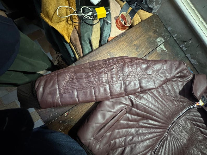 May include: A burgundy leather jacket with quilted stitching and a ribbed cuff. The jacket features the word "AMOR" stitched on the sleeve. The jacket has a front pocket and a zipper closure.