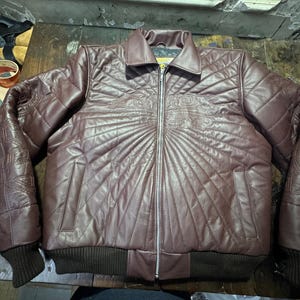 May include: A maroon leather jacket with a quilted design and a zipper closure. The jacket has a collar, two pockets, and ribbed cuffs and hem. Decorative stitching is visible on the shoulders and chest.