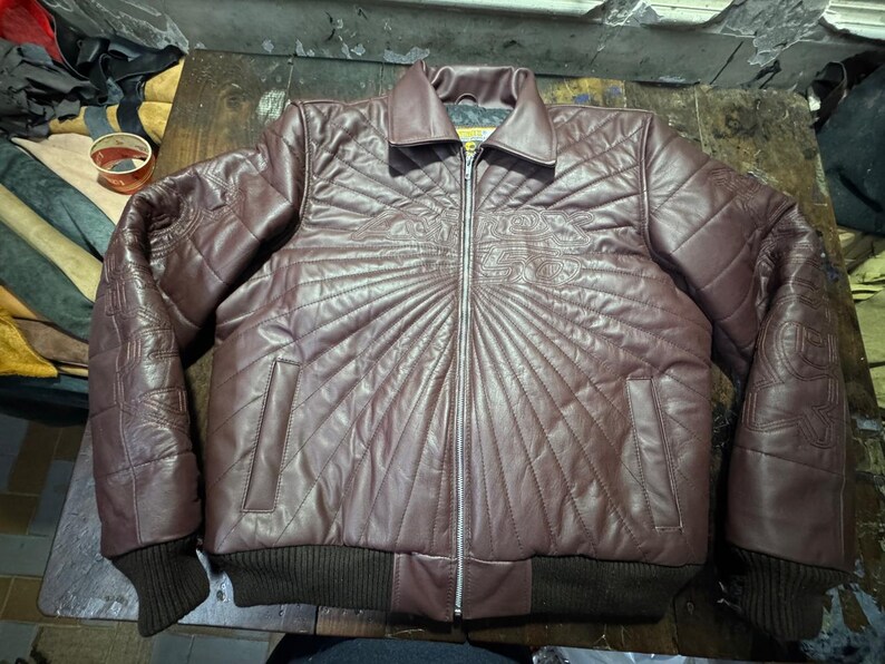 May include: A maroon leather jacket with a quilted design and a zipper closure. The jacket has a collar, two pockets, and ribbed cuffs and hem. Decorative stitching is visible on the shoulders and chest.