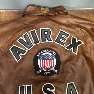 May include: Brown leather jacket with the word "AVIREX" in white block letters. Below the word "AVIREX" is a patch with the words "ALL AMERICANS ESTABLISHED 1975" and the USA flag. The word "USA" is below the patch.