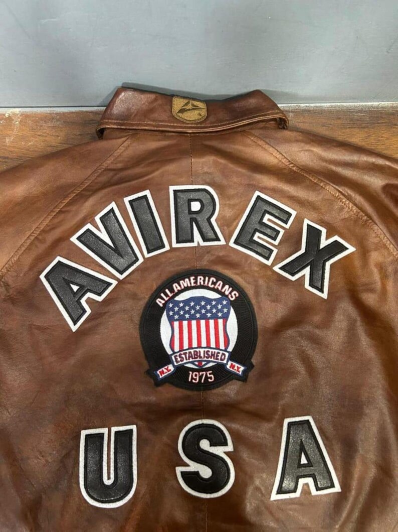 May include: Brown leather jacket with the word "AVIREX" in white block letters. Below the word "AVIREX" is a patch with the words "ALL AMERICANS ESTABLISHED 1975" and the USA flag. The word "USA" is below the patch.