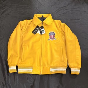 May include: A vibrant yellow bomber jacket with a zip, a collar, and a chest patch. The jacket has white and yellow striped cuffs and hem. The patch displays a red, white, and blue design and the word "AVIREX".