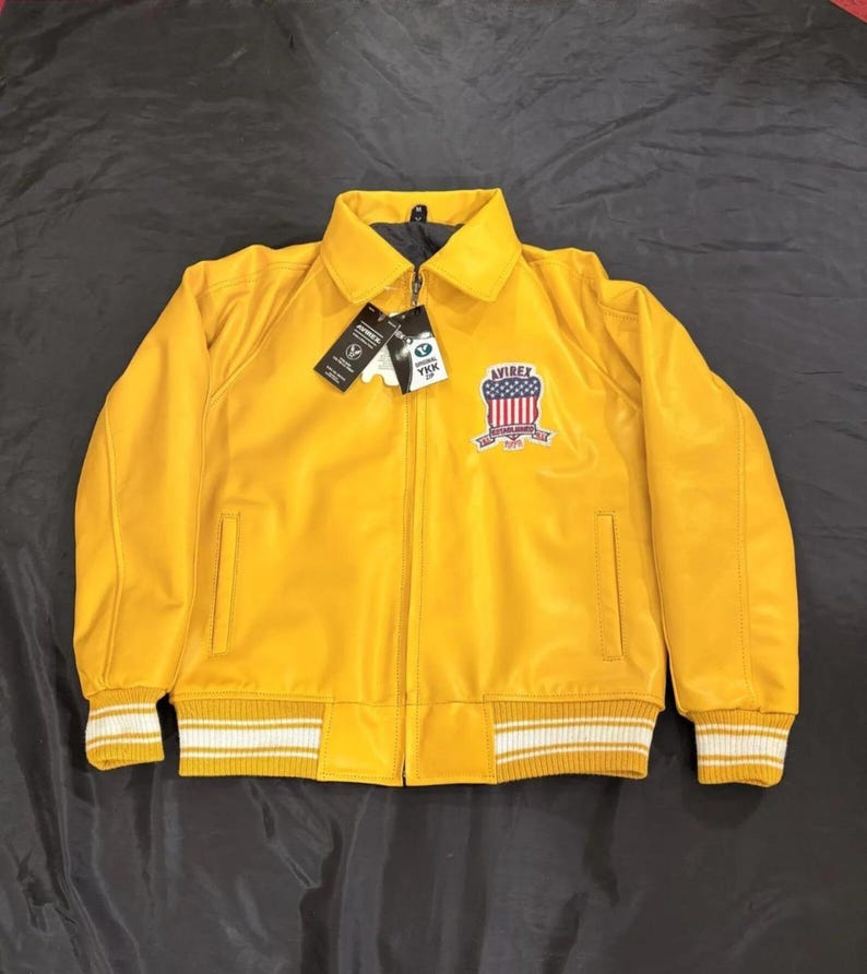May include: A vibrant yellow bomber jacket with a zip, a collar, and a chest patch. The jacket has white and yellow striped cuffs and hem. The patch displays a red, white, and blue design and the word "AVIREX".