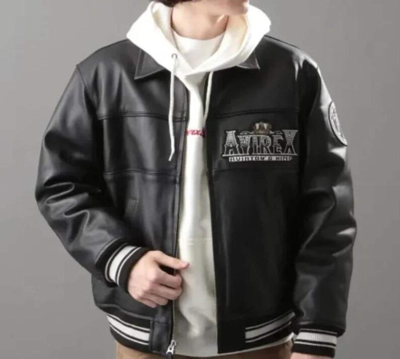 May include: Black leather jacket with a white hooded sweatshirt underneath. The jacket features a collar, a zipper closure, and striped cuffs and hem. The jacket has the text "AVIREX" on the chest.