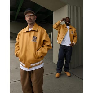 May include: Two people wearing mustard yellow leather jackets with a white and cream striped hem. The jackets have a patch on the chest. One person wears a black cap and glasses, the other wears a white t-shirt and dark blue jeans.