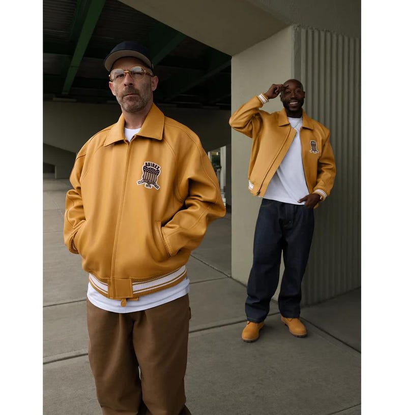 May include: Two people wearing mustard yellow leather jackets with a white and cream striped hem. The jackets have a patch on the chest. One person wears a black cap and glasses, the other wears a white t-shirt and dark blue jeans.