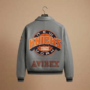 Puede incluir: Una chaqueta universitaria de cuero gris con un gran logotipo circular de los New York Knicks en la espalda, bordado en naranja y azul marino. La palabra AVIREX est&aacute; bordada en naranja debajo del logo. La chaqueta tiene cuello y pu&ntilde;os acanalados.