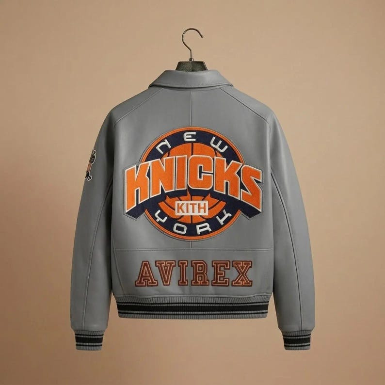 Puede incluir: Una chaqueta universitaria de cuero gris con un gran logotipo circular de los New York Knicks en la espalda, bordado en naranja y azul marino. La palabra AVIREX est&aacute; bordada en naranja debajo del logo. La chaqueta tiene cuello y pu&ntilde;os acanalados.