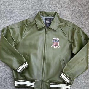 May include: Olive green leather jacket with a zip-up front, collar, and ribbed cuffs and hem. Features a patch with an American flag design and the brand name "AVIREX". The cuffs and hem have white stripes.
