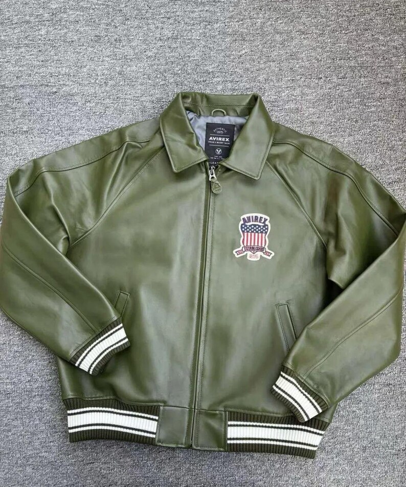 May include: Olive green leather jacket with a zip-up front, collar, and ribbed cuffs and hem. Features a patch with an American flag design and the brand name "AVIREX". The cuffs and hem have white stripes.