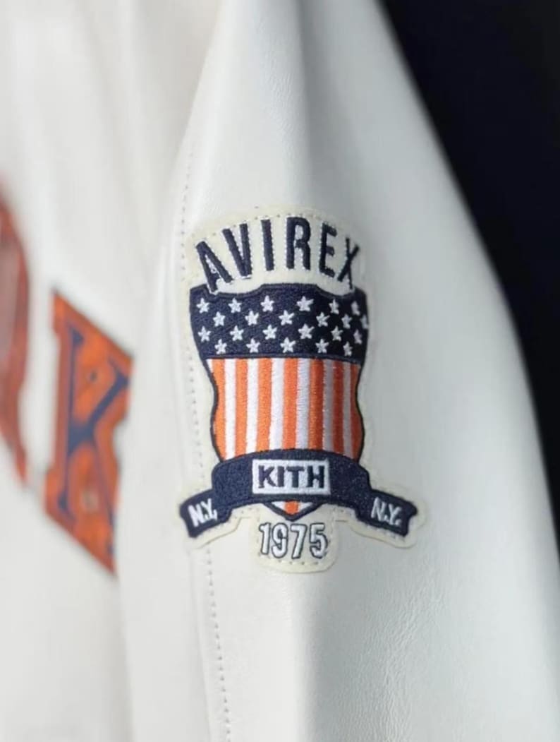 Pode incluir: Close-up de uma jaqueta de couro branca com um patch. O patch apresenta a palavra "AVIREX" acima de um desenho da bandeira americana. Abaixo, a palavra "KITH" e o ano "1975".
