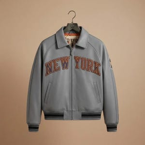 Puede incluir: Una chaqueta gris con cremallera, cuello cl&aacute;sico y pu&ntilde;os y dobladillo acanalados. La parte delantera muestra el texto NEW YORK en letras negritas con bordes oscuros. El forro interior es visible en el cuello con un dise&ntilde;o estampado.