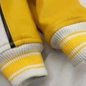 May include: Close-up of a yellow and white varsity jacket sleeve. The sleeve features a ribbed cuff with yellow and white stripes. The jacket has a yellow body and a white panel with a black stripe.