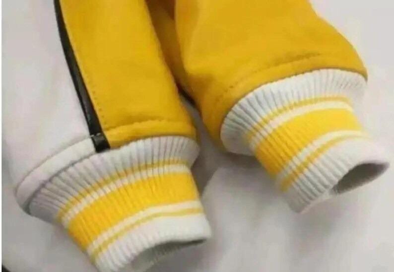 May include: Close-up of a yellow and white varsity jacket sleeve. The sleeve features a ribbed cuff with yellow and white stripes. The jacket has a yellow body and a white panel with a black stripe.
