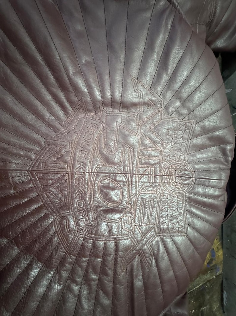 May include: A burgundy leather jacket with a quilted design. The back of the jacket features an embroidered emblem with the words "SOLDIER KICKOFF" and star details. The stitching creates a sunburst pattern.