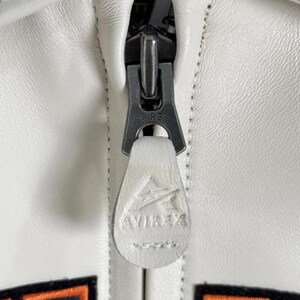 Pode incluir: Close-up de um z&iacute;per de jaqueta de couro branco com o nome da marca "AVIREX" vis&iacute;vel no puxador do z&iacute;per. O z&iacute;per &eacute; prateado e a jaqueta tem costuras brancas.