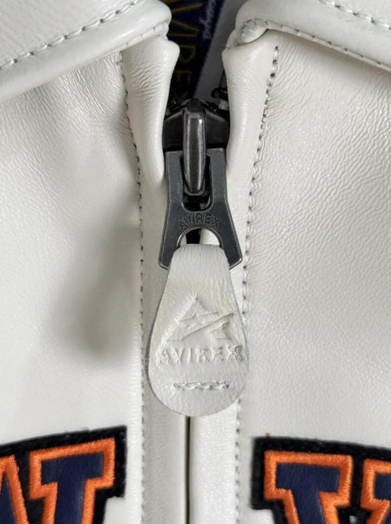 Pode incluir: Close-up de um z&iacute;per de jaqueta de couro branco com o nome da marca "AVIREX" vis&iacute;vel no puxador do z&iacute;per. O z&iacute;per &eacute; prateado e a jaqueta tem costuras brancas.