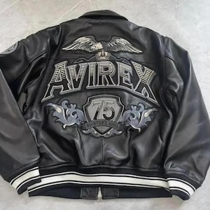 May include: Black leather jacket with white and grey embroidered details. The back features the word "AVIREX" above a shield with the number "75" and the text "AVIATOR'S KING". The jacket has a white and black striped trim.