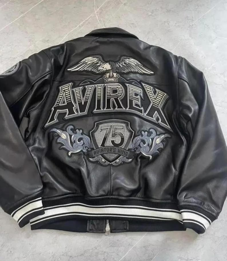 May include: Black leather jacket with white and grey embroidered details. The back features the word "AVIREX" above a shield with the number "75" and the text "AVIATOR'S KING". The jacket has a white and black striped trim.