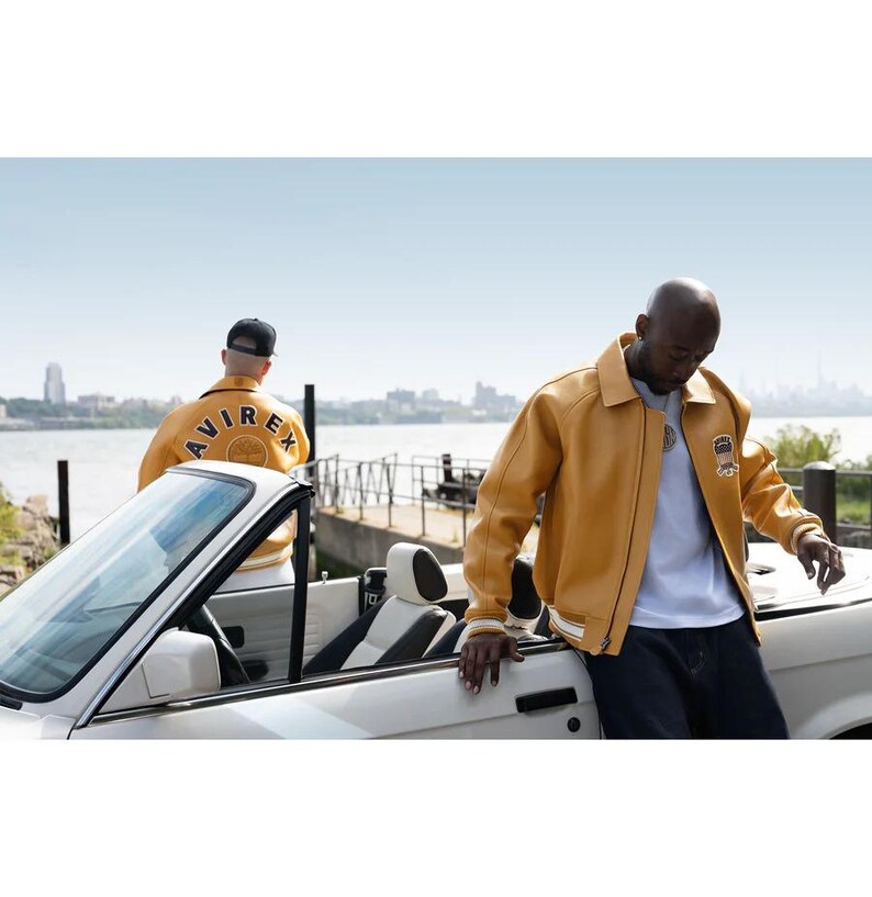 May include: Mustard yellow leather jacket with white trim and a small chest patch. A person wears the jacket next to a white convertible car. Another person wears a similar jacket with "AVIREX" on the back. The jackets are a classic style.